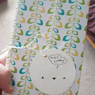 A photograph of a rectangular gift wrapped in paper with a design resembling leaves on plant stems in yellow, green and blue, with a white, circular gift tag in the middle. The tag has a smiling small robot style face drawn on it and a speech bubble that reads "happy birthday!".