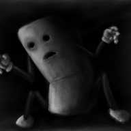 A vaguely rectangular robot with jointed arms and legs, bent into the darkened corner of a room, looming out of the shadows, hands outstretched. Its eyes are empty, bored holes in its surface and its mouth a gaping, down-turned slash.